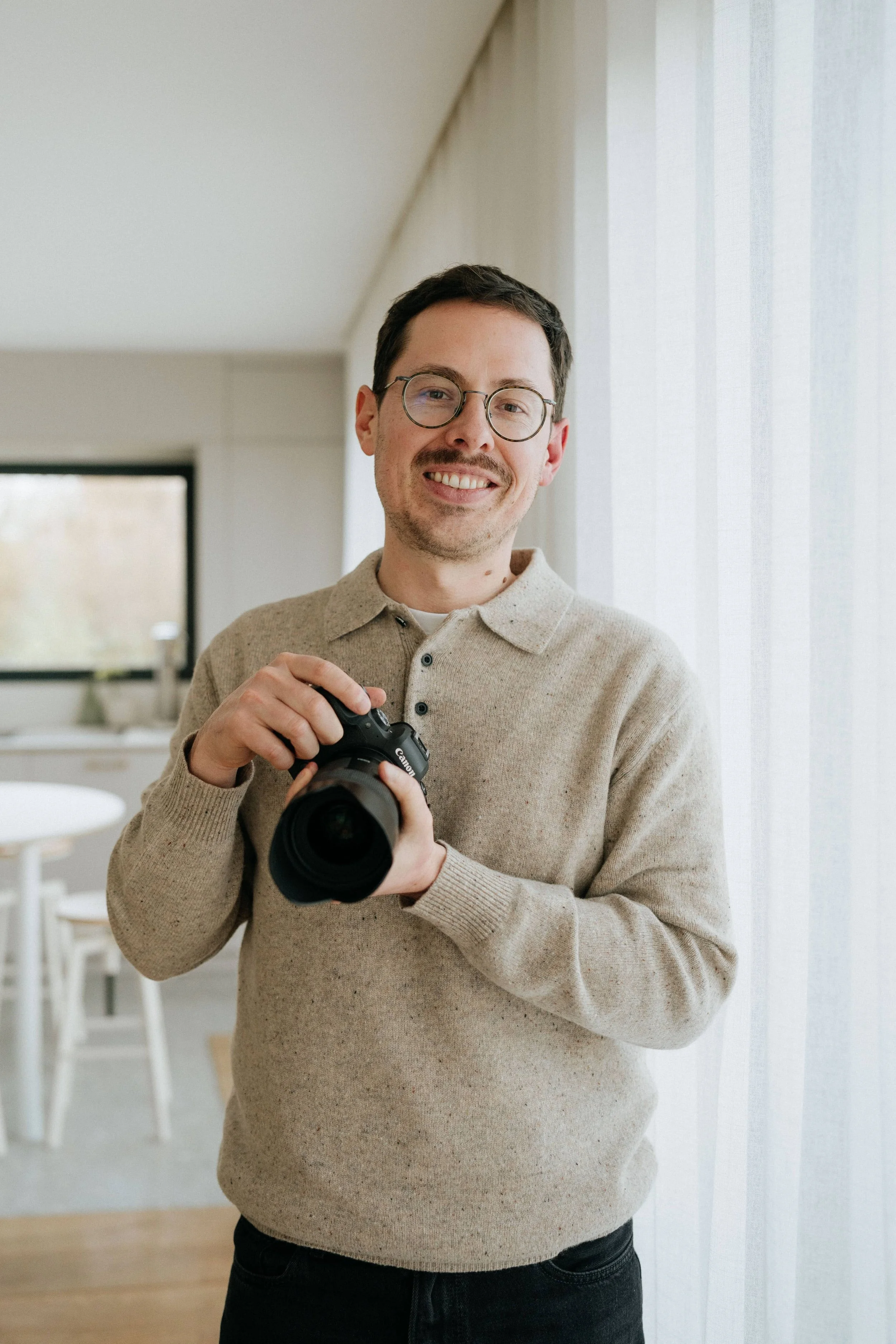Portrait du photographe Max De Hulster. Il se tien debout tout en souriant et en tenant son appareil photo dans les mains.