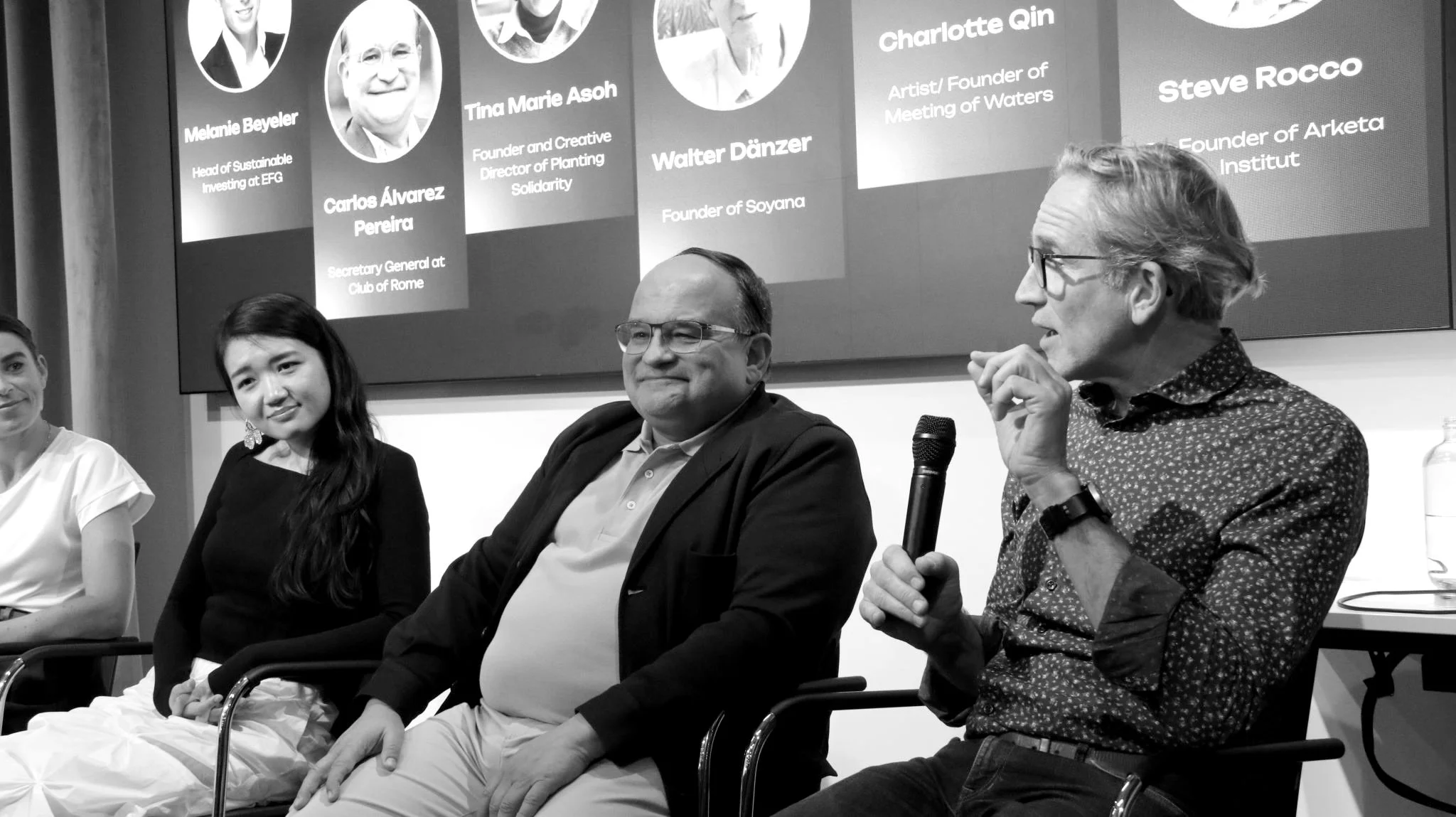 Steve (a man in glasses and a dark button down shirt) speaks into a microphone at a panel discussion as the other panelists look on.