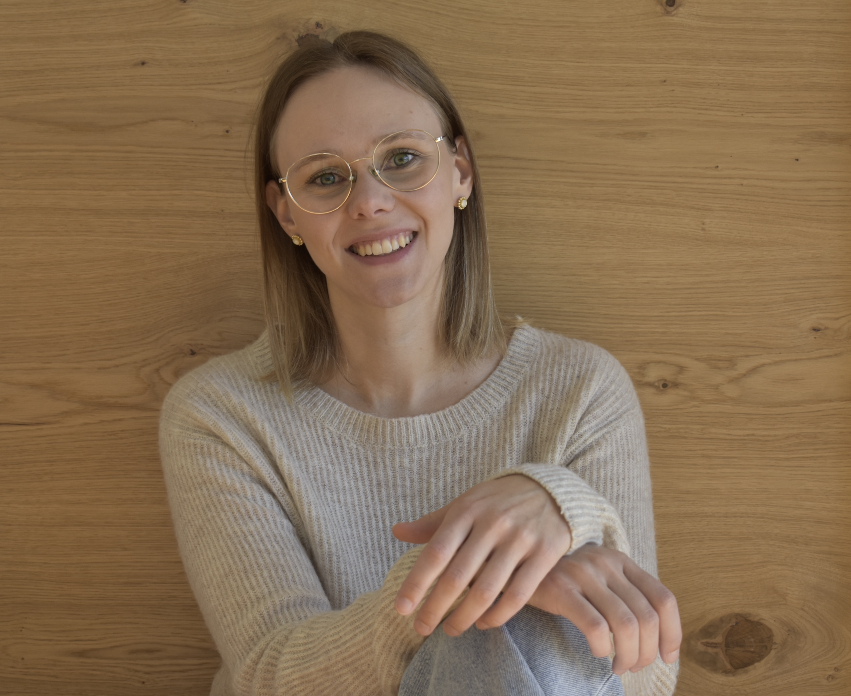 Eine lächelnde Frau mit Brille und Ohrringen sitzt vor einer Holzwand, trägt einen cremefarbenen Pullover und hält ihre Beine mit den Armen umarmt.