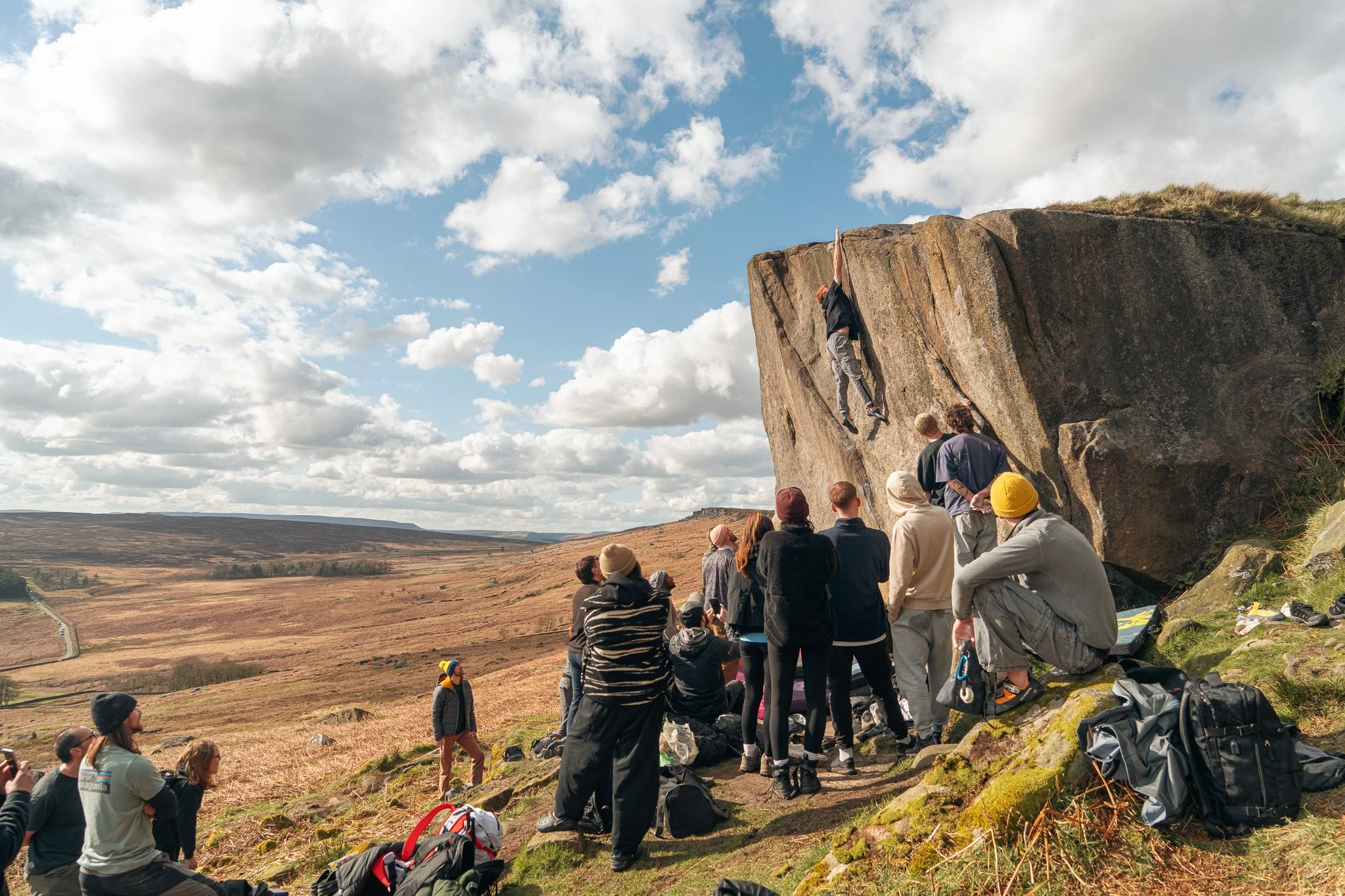 A group of people watching a rock climber on a boulder in a scenic outdoor landscape under a cloudy sky, with backpacks and gear on the ground.