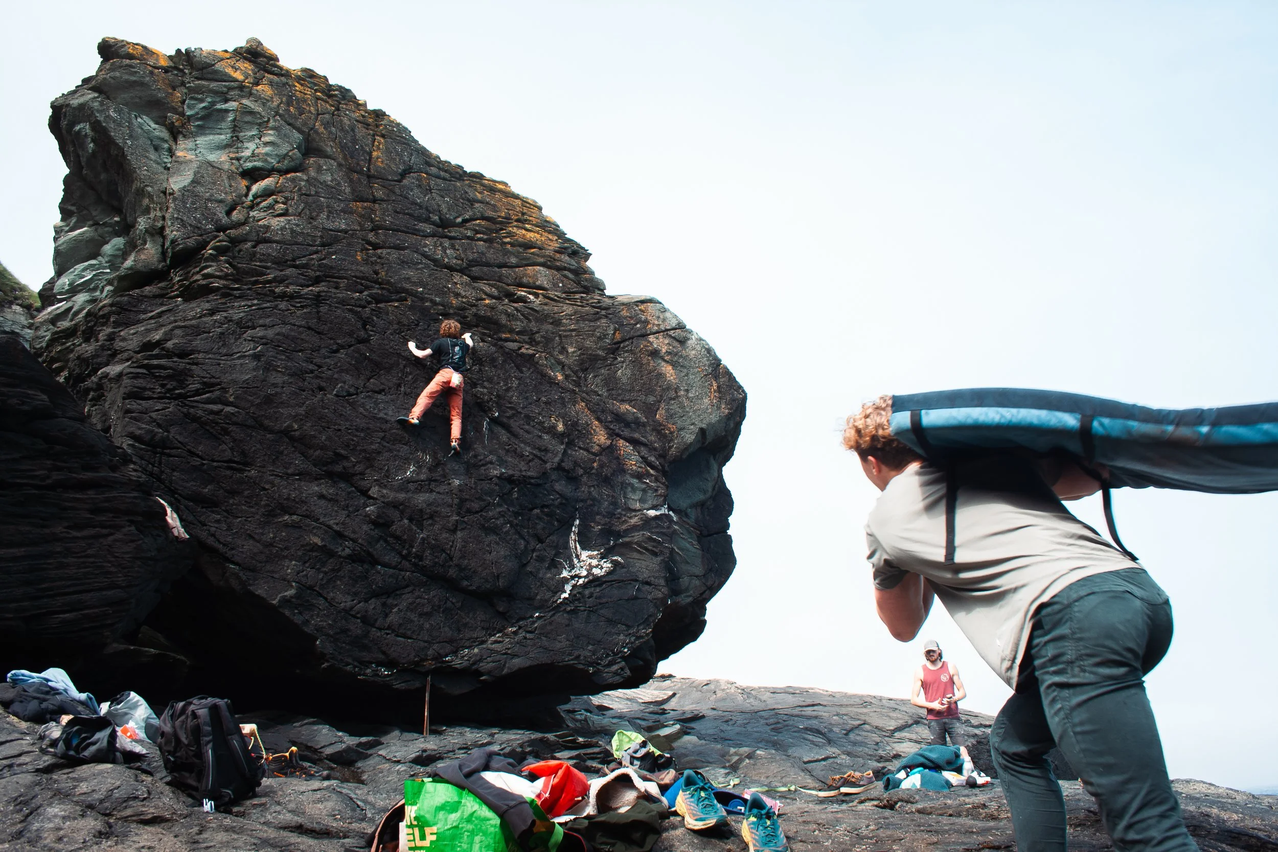 Person bouldering on a large rock with another person carrying a crash pad, and equipment scattered on the ground.
