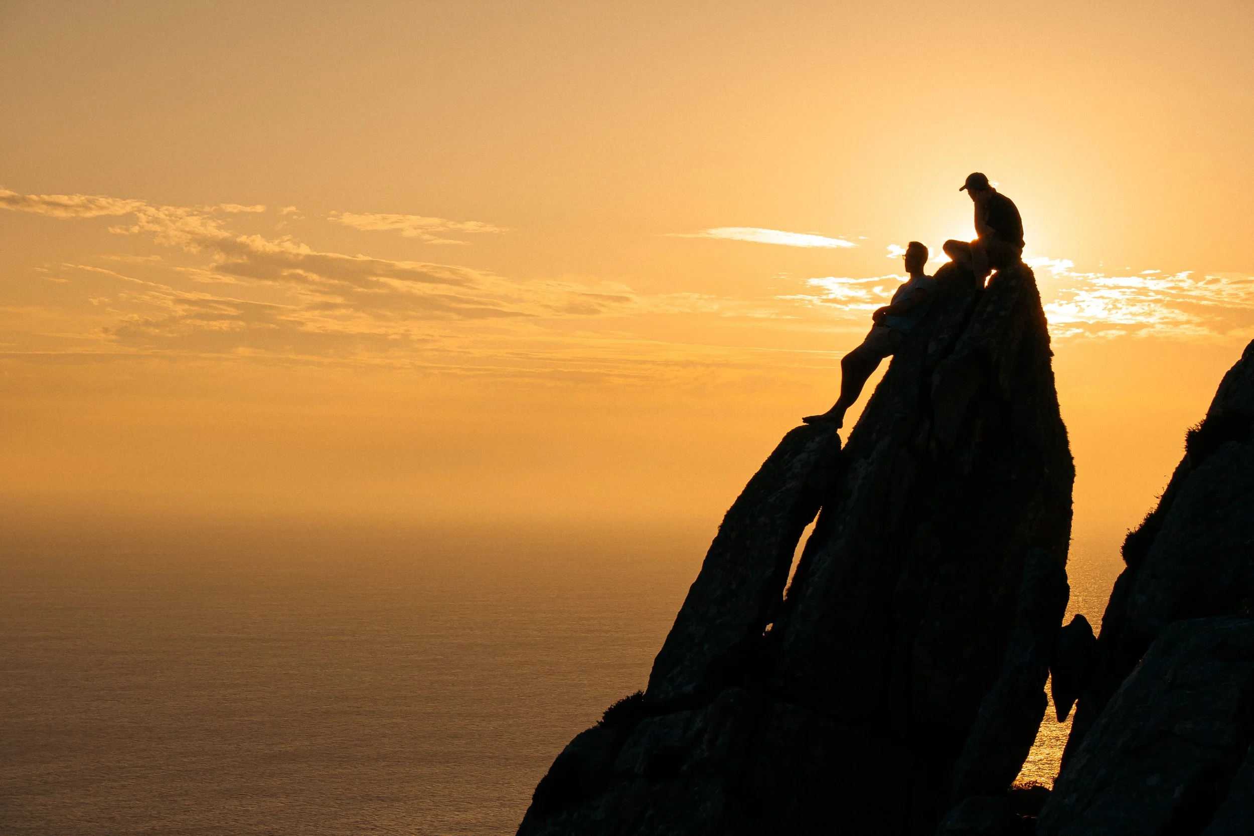 Silhouette of two people sitting on a rocky peak during sunset with an ocean view.