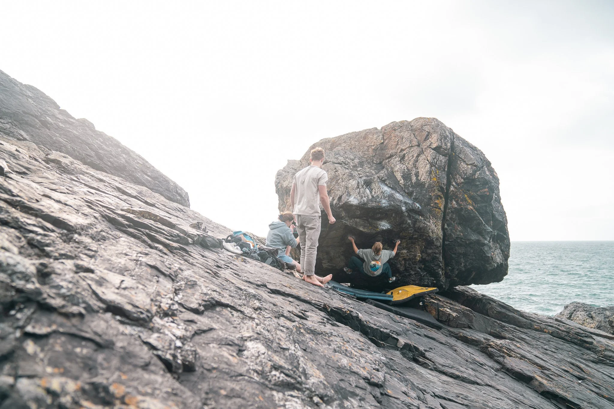 Person bouldering on rocky coastline with two observers and gear nearby, ocean in background.