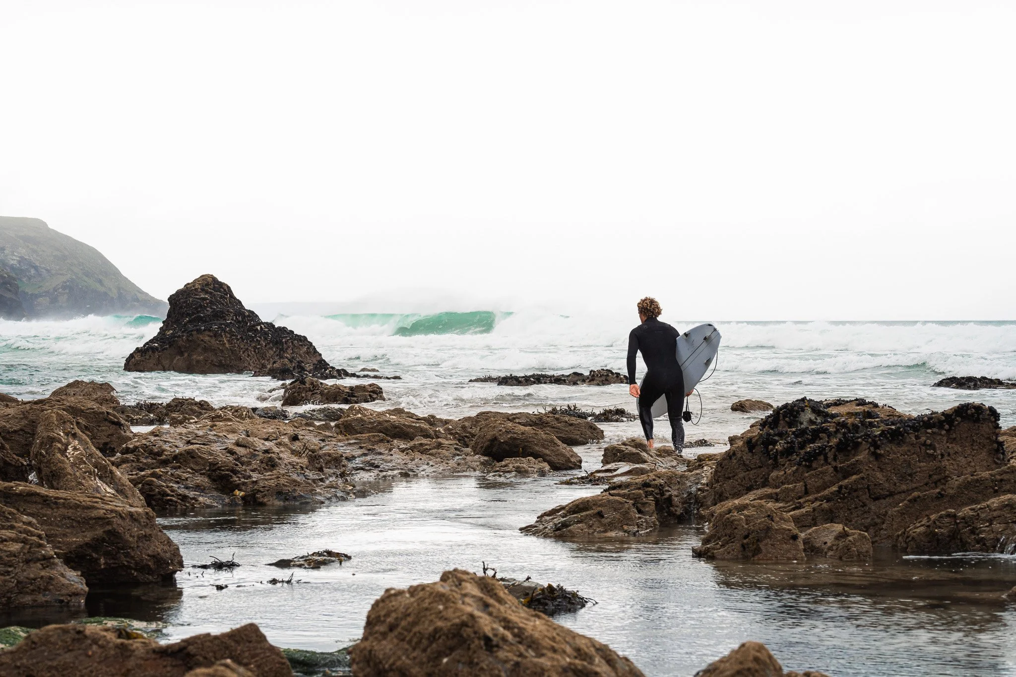 Surfer walking on rocky shoreline towards ocean waves.