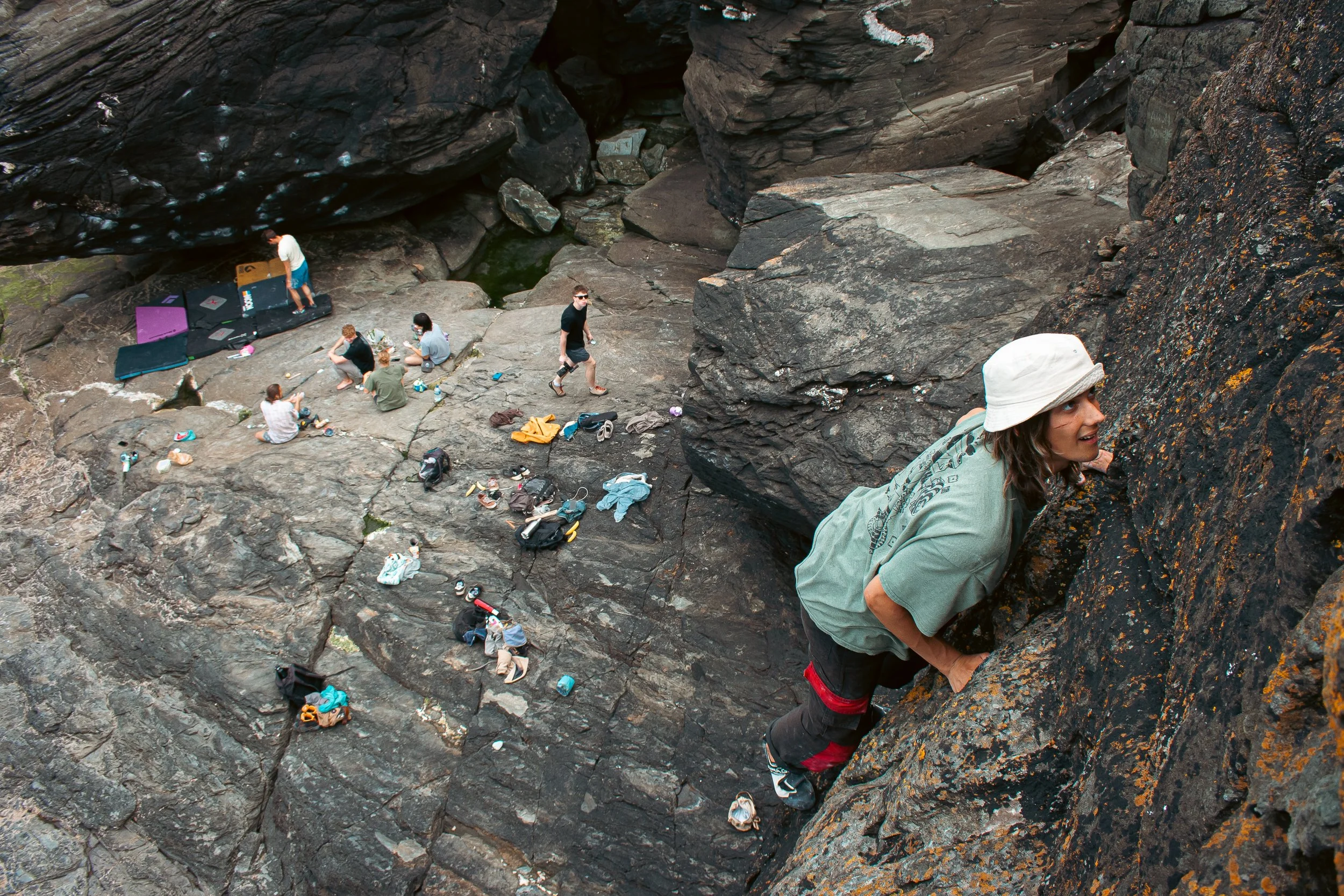 Person climbing a rock face wearing a white bucket hat, with multiple people sitting and standing on rocky ground nearby, surrounded by climbing gear and bags.