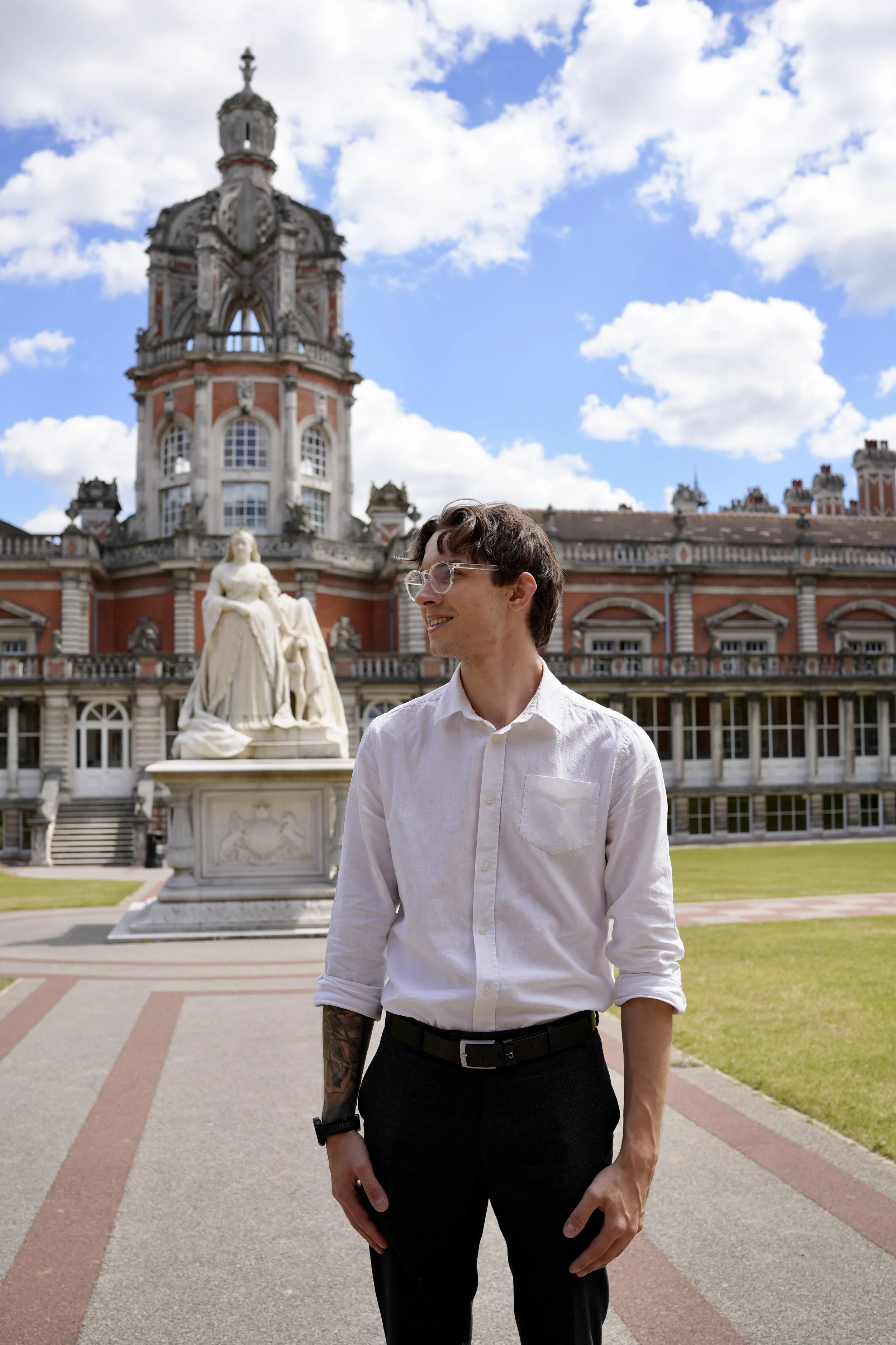 A young man with glasses, wearing a white shirt and black pants, standing outdoors in front of a historic castle with a statue in the foreground and a partly cloudy sky.