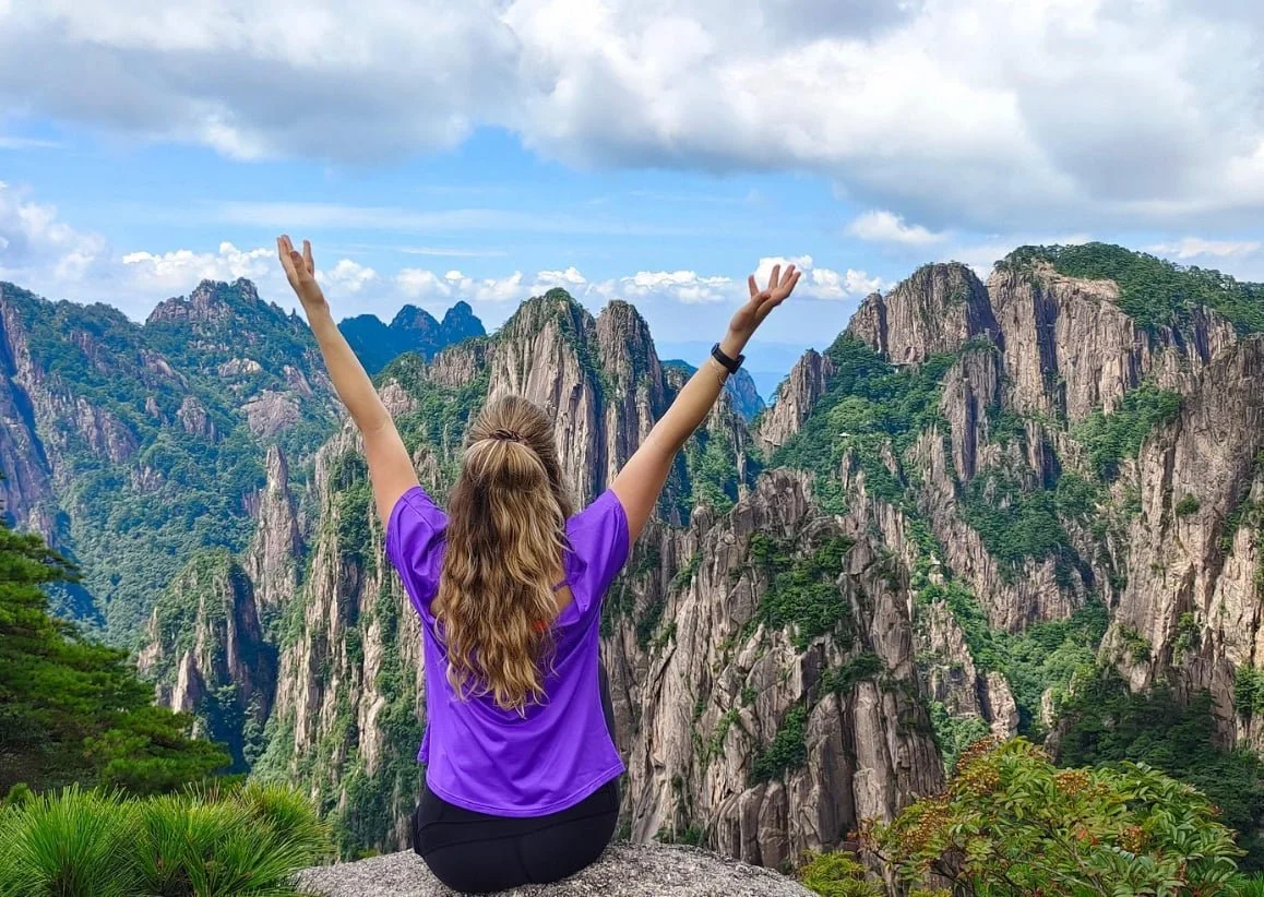 A woman sitting on a rock with her arms raised in front of a mountainous landscape with green trees and blue sky.