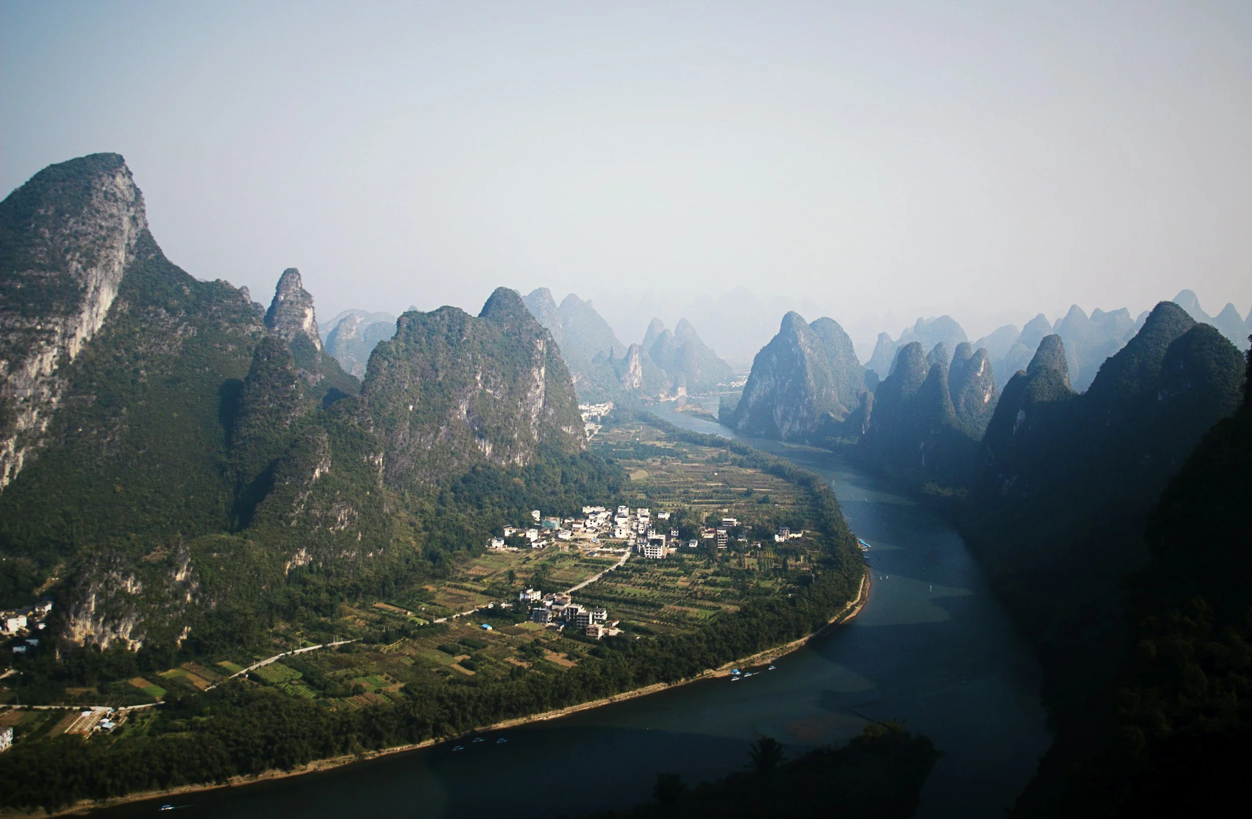 Scenic landscape of tall, green, jagged mountains surrounding a winding river with small buildings and farmland at their base.