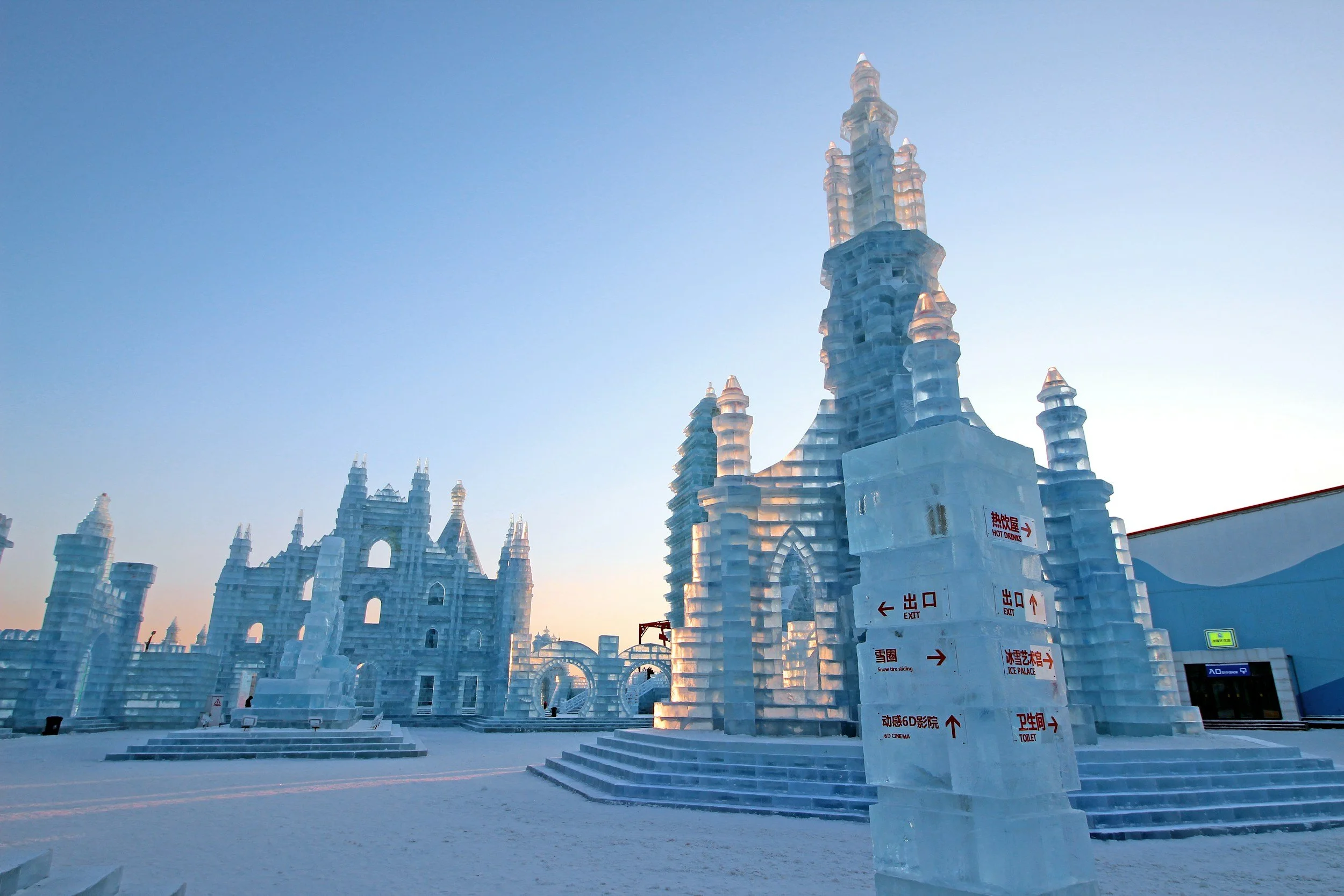 Ice castle structures resembling castles with towers and spires, set against a pale blue sky at sunrise or sunset, with signs in Chinese and English near the entrance.