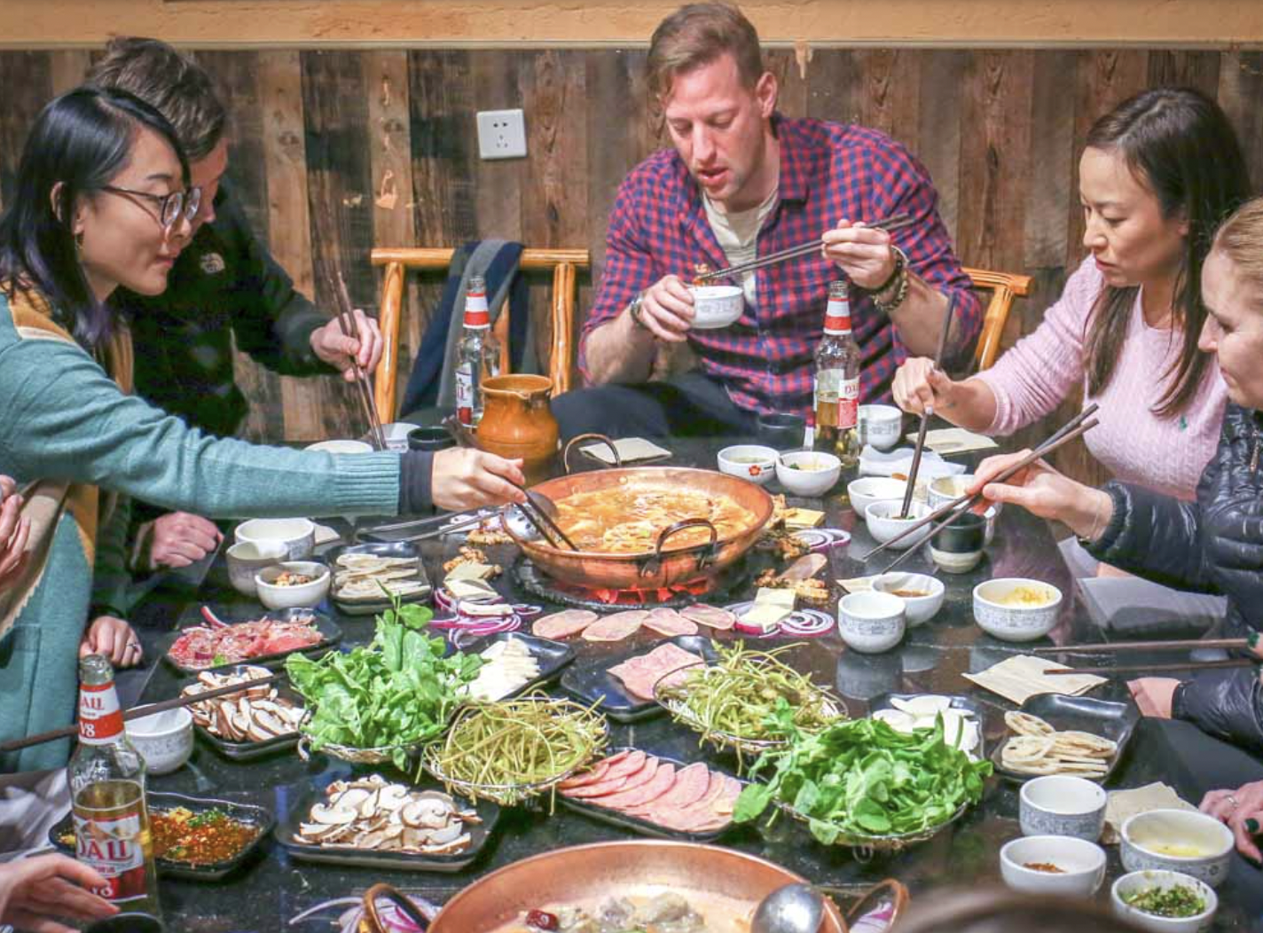 A group of people enjoying a hot pot meal at a restaurant table with various vegetables, meats, and condiments, using chopsticks.