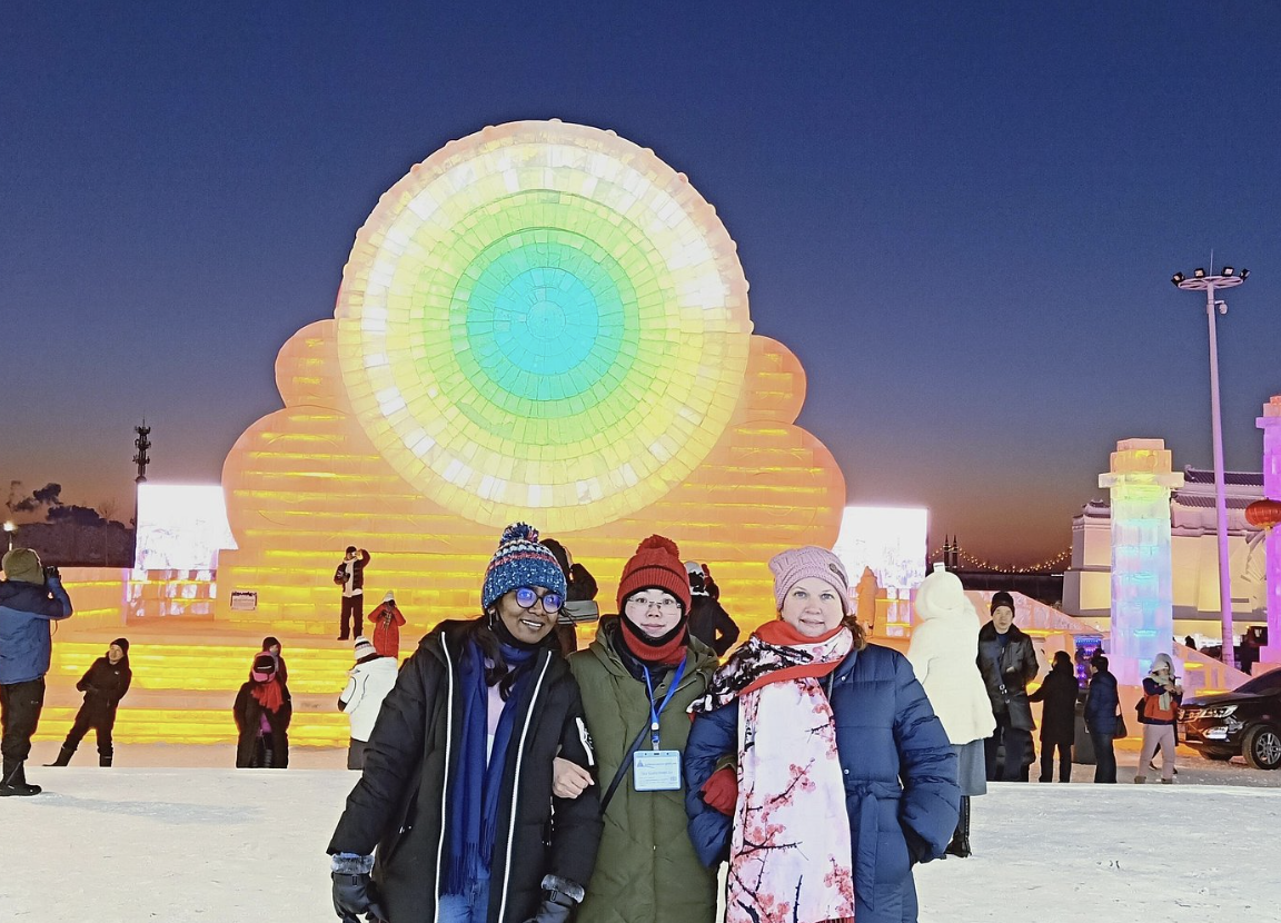 People dressed warmly standing in front of an illuminated colorful ice sculpture of a large, intricate circular design at an outdoor winter event during dusk.