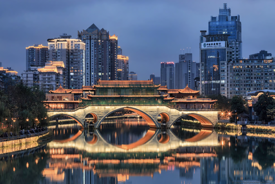 City skyline with a traditional Chinese-style bridge over a river, illuminated at night with reflections in the water.