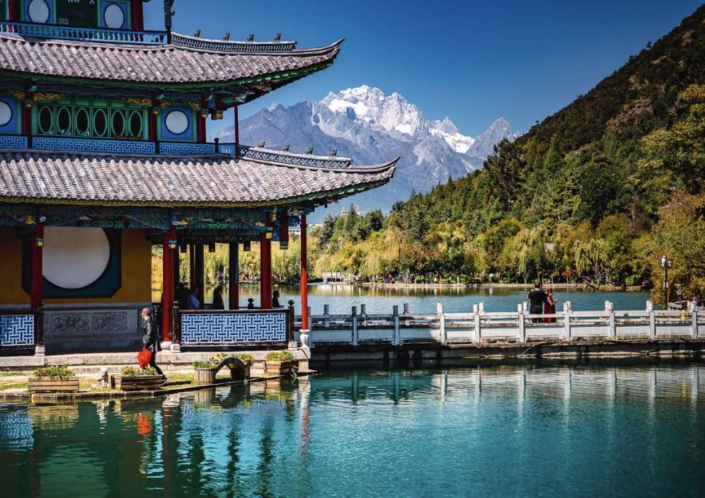 A traditional Chinese pavilion over a lake with mountains in the background, colorful trees, and people walking on the bridge.