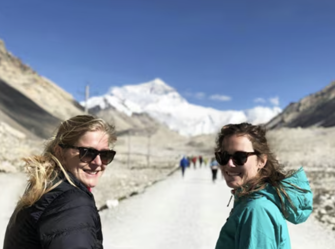 Two smiling women wearing sunglasses and jackets standing on a trail in a mountainous snowy landscape with snow-capped peaks in the background.