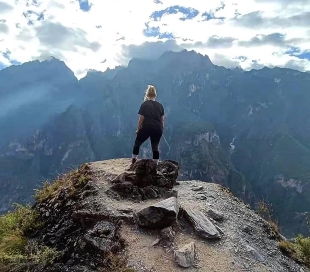 A person standing on a rocky outcrop overlooking a mountain range with cloudy skies.