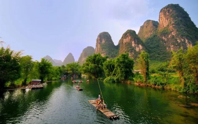 A river scene with green trees and rocky mountains in the background, with people on bamboo rafts floating on the water.