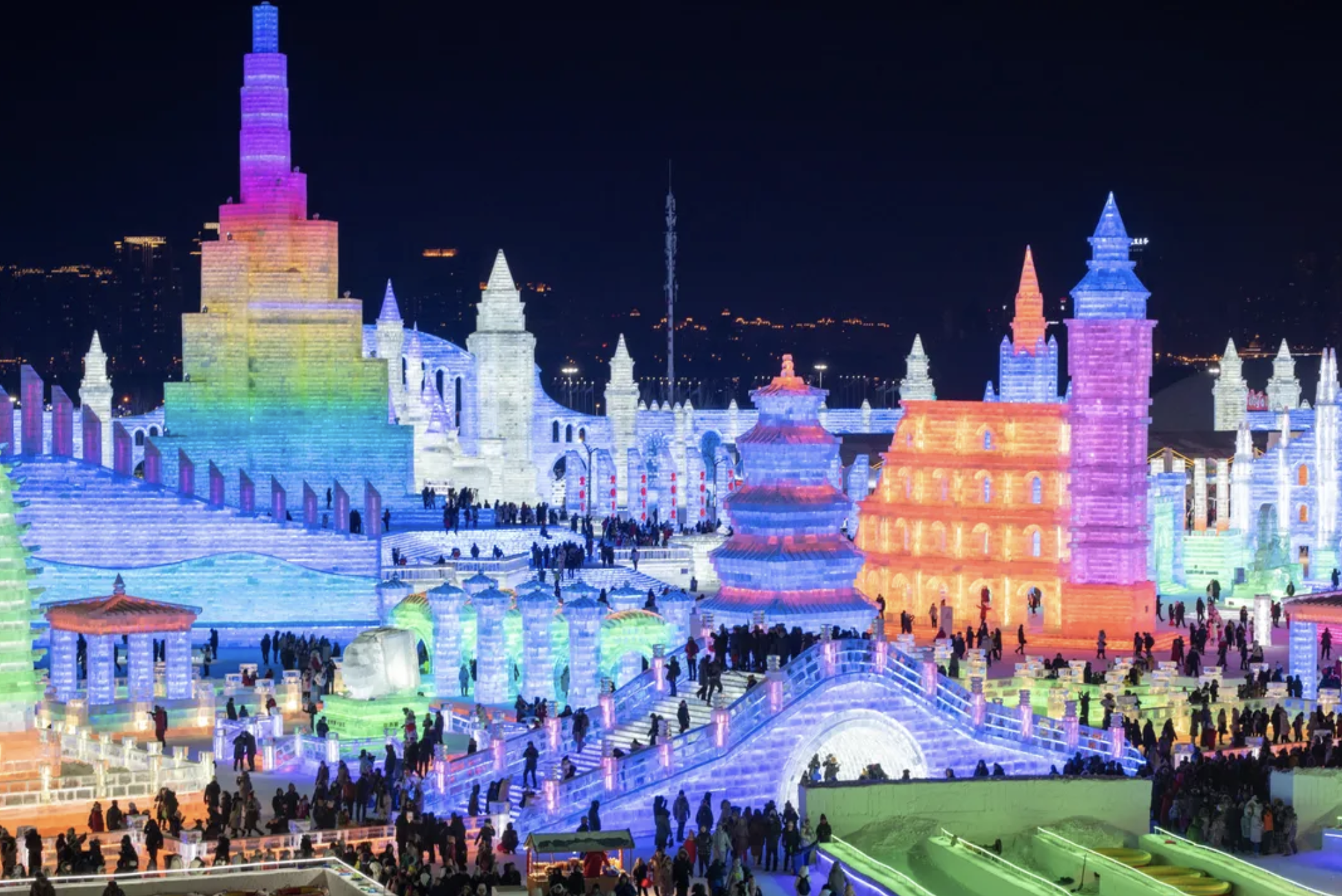 Colorfully illuminated ice castle at night with numerous visitors walking around.