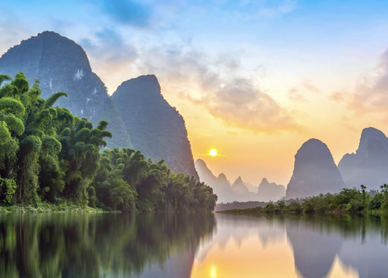 A serene landscape featuring a river with a reflection of green trees and towering karst mountains at sunrise or sunset with a partly cloudy sky.