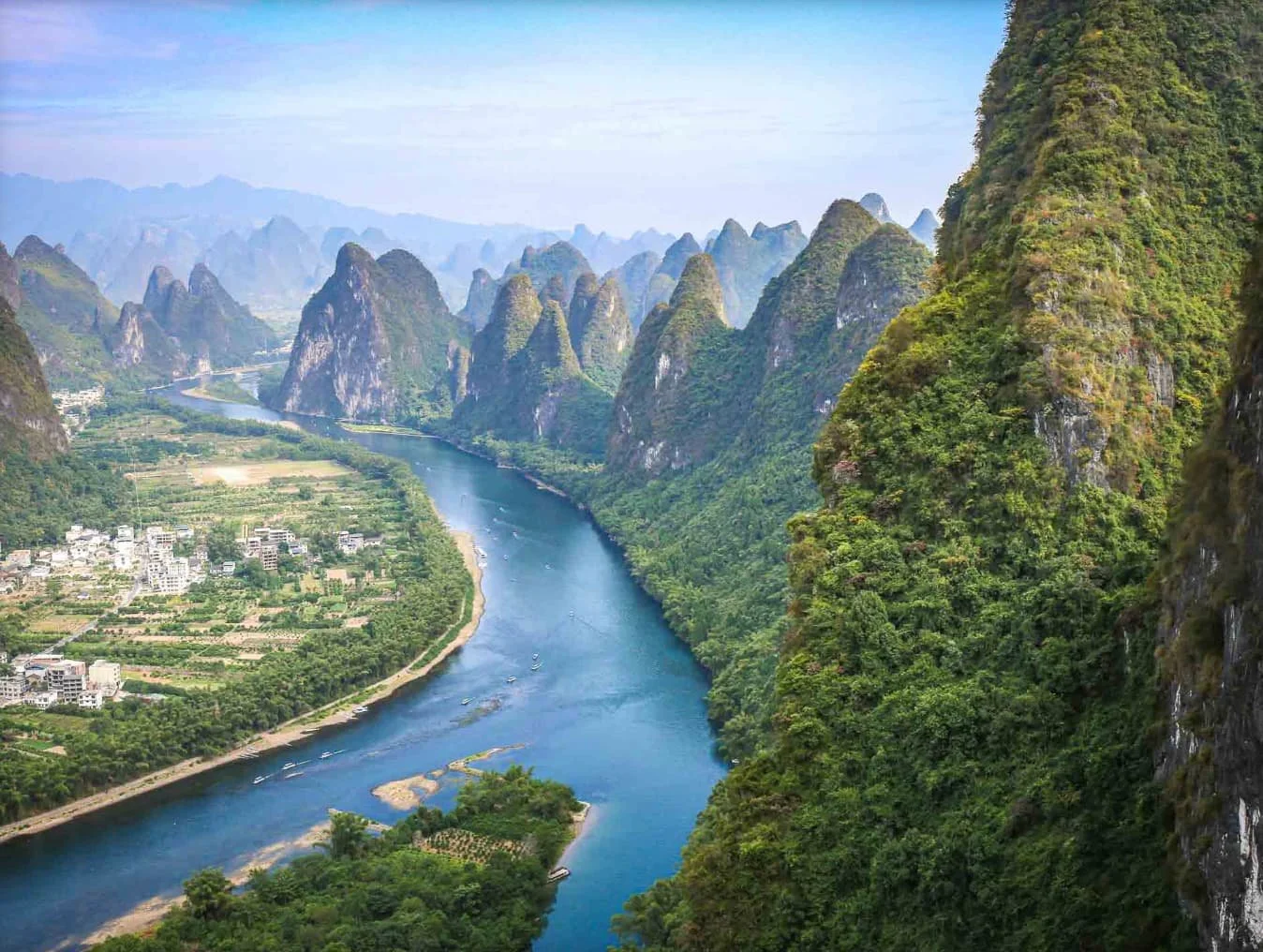 A scenic view of a winding river flowing through lush green mountains with small buildings and farmland on the riverbank.