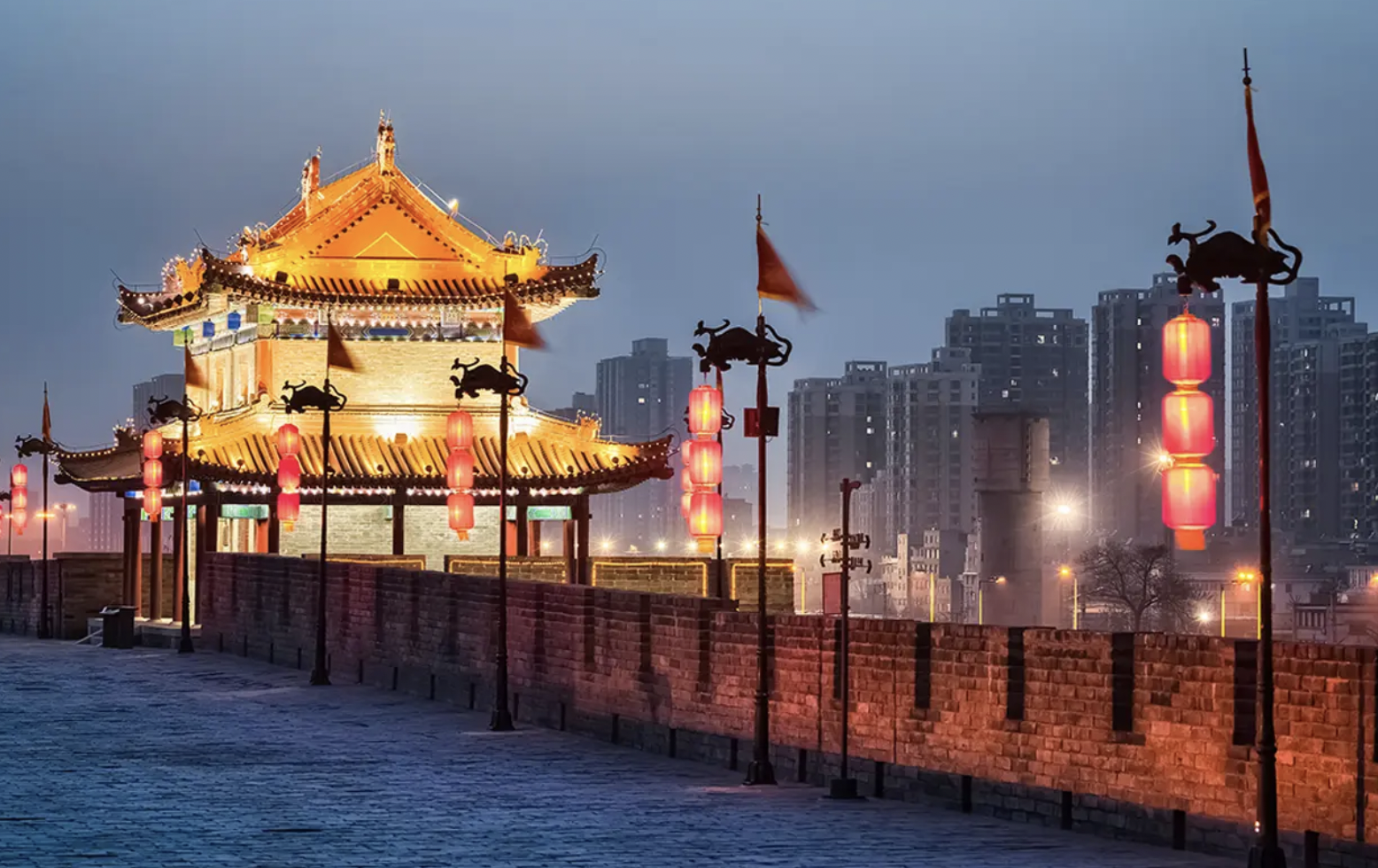 Traditional Chinese pavilion illuminated at night along a pathway with red lanterns and flags, background of modern high-rise buildings.