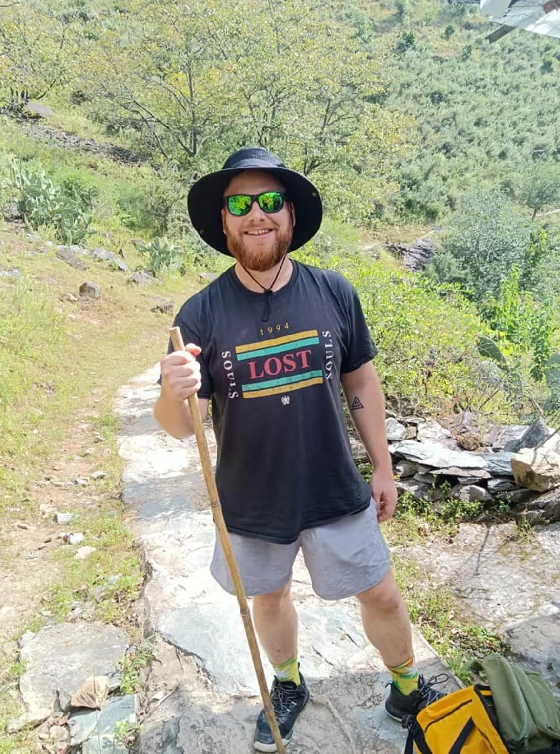 A man with a beard wearing a wide-brimmed hat, sunglasses with reflective green lenses, a black T-shirt that says 'LOST' and light-colored shorts, standing on a rocky trail in a lush, green outdoor setting, holding a walking stick and smiling.