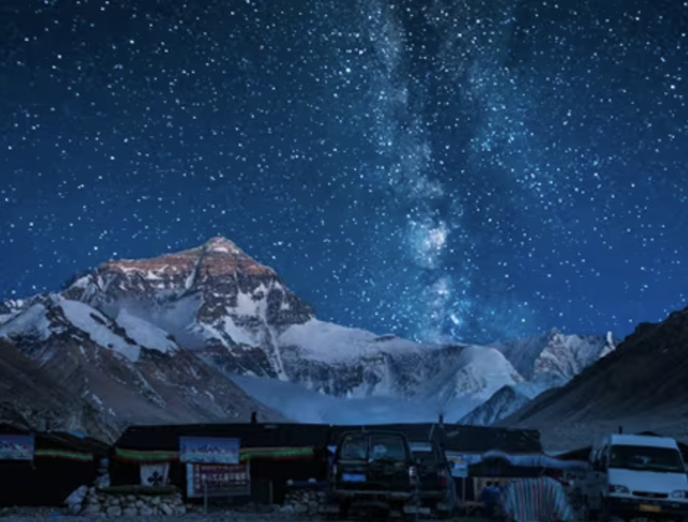 Snow-capped mountains under a starry night sky with the Milky Way, with parked cars and buildings in the foreground.