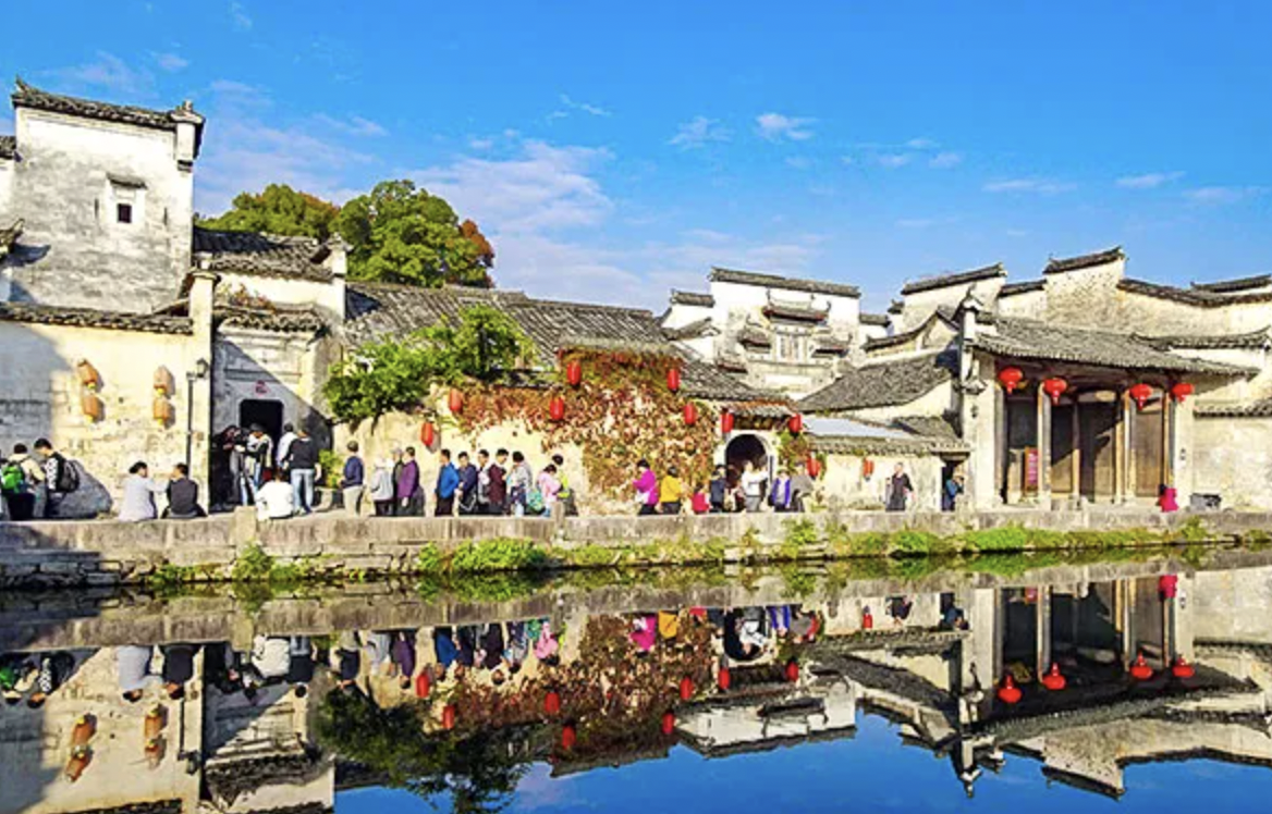 Traditional Chinese buildings with red lanterns along a canal, reflecting in the water, with people walking and gathering outside under a blue sky.