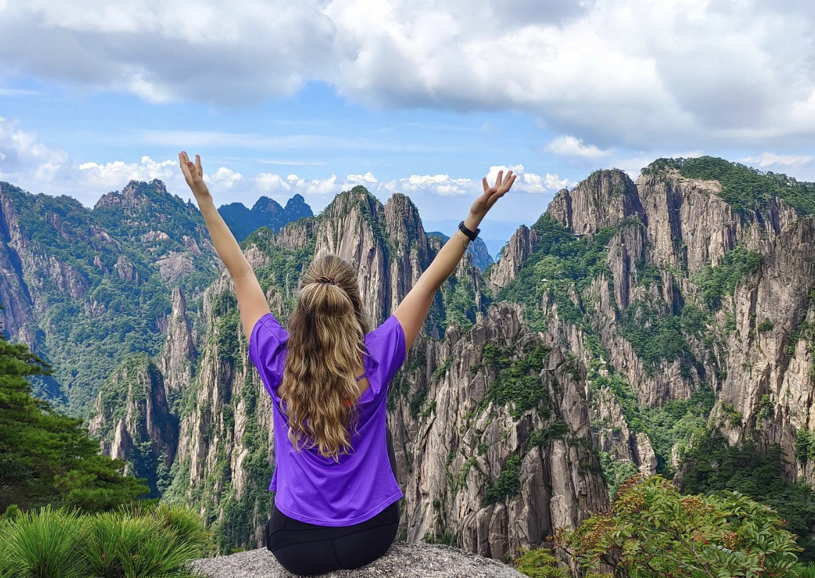 A woman with long blond hair sitting on a rock with her arms raised, overlooking a mountain landscape with rugged, green peaks and a partly cloudy sky.