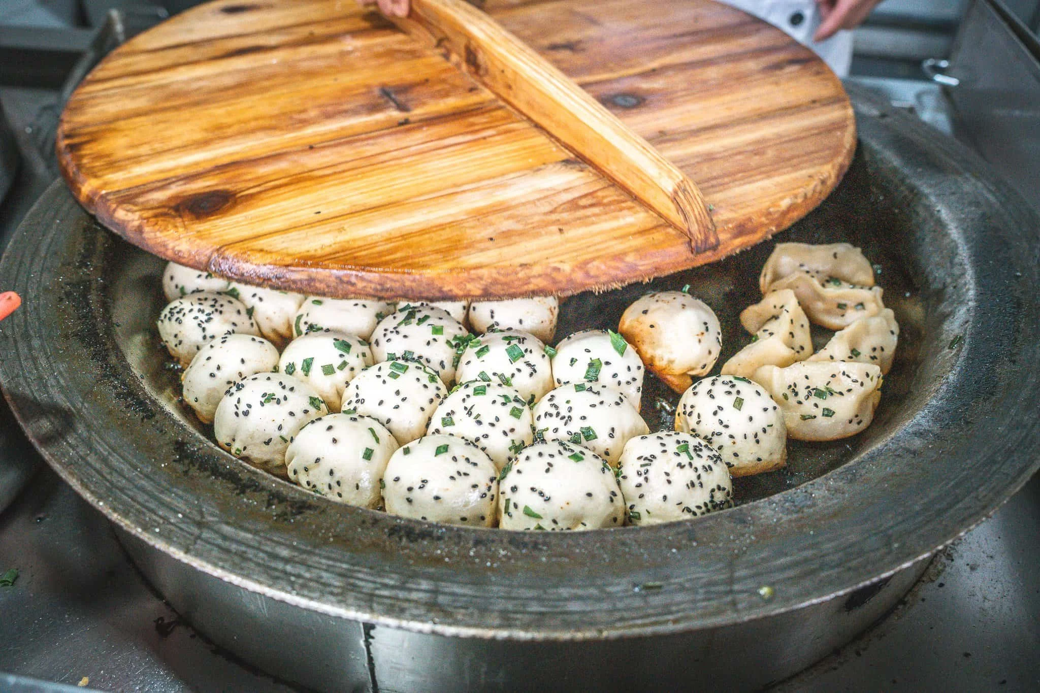 Dumplings cooking on a stovetop with a wooden lid partially covering them, garnished with chopped green onions and black sesame seeds.