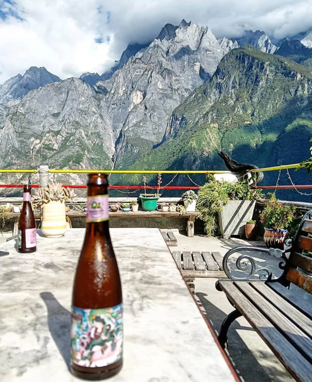 Outdoor patio with wooden benches, potted plants, and bottles on a table, overlooking a mountainous landscape with rocky peaks and green slopes under partly cloudy skies.