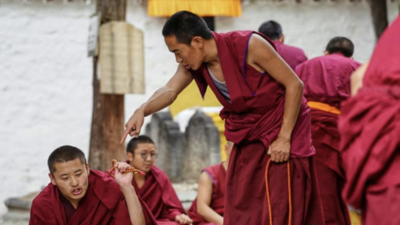 A monk in a red robe bending over to speak to a seated young monk holding prayer beads, with other monks in the background.