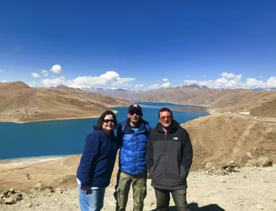 Three people standing together outdoors against a backdrop of a large lake and brown, hilly terrain under a clear blue sky.