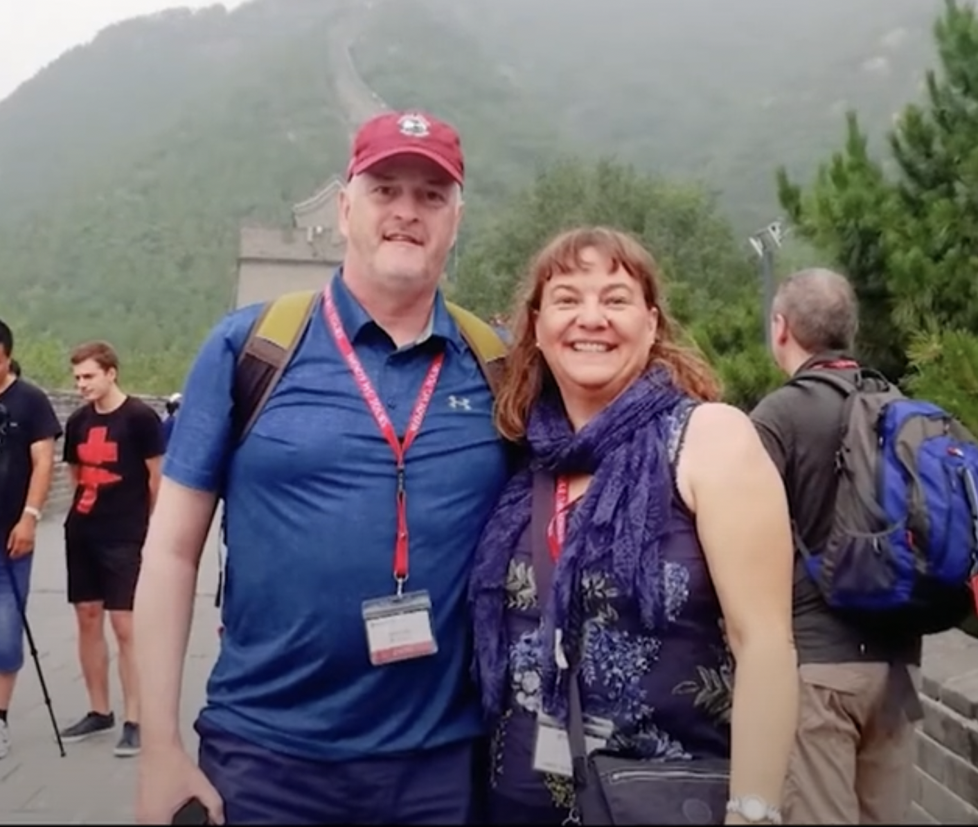 Two people smiling outdoors with a mountain structure and greenery in the background. The man on the left is wearing a blue shirt and a red cap, and the woman on the right is wearing a purple scarf and a floral top, with people in the background.