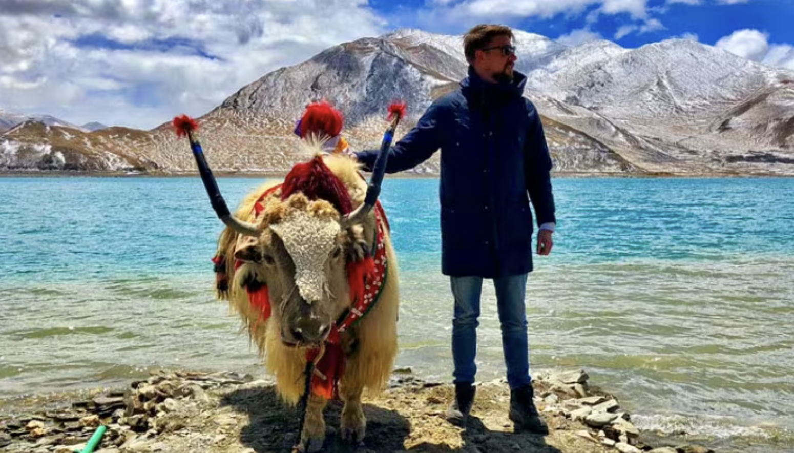 A man standing on a rocky shore with a decorated yak, a mountainous landscape with snow-capped peaks in the background, and a blue lake.