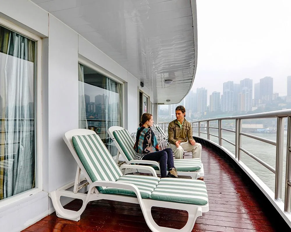 Two people sitting on lounge chairs with green and white stripes on a cruise ship balcony, overlooking city skyline and river.
