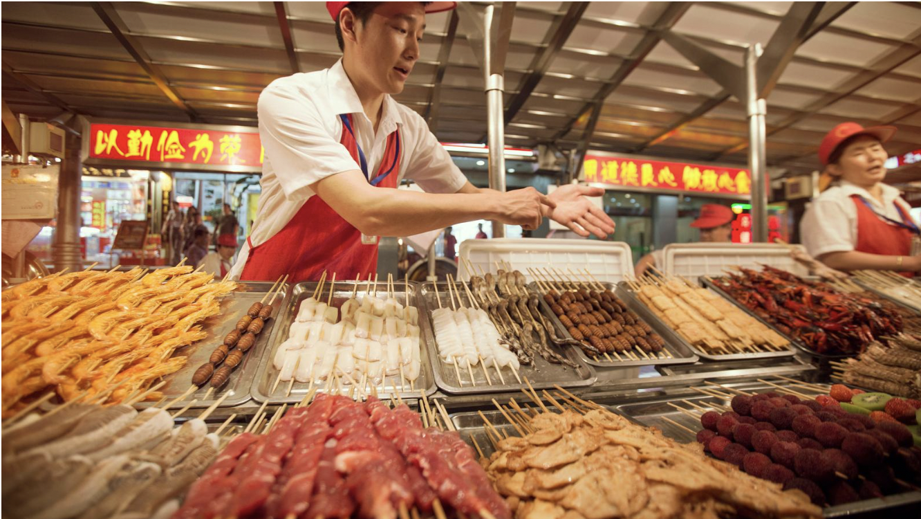 A person interacting with a food vendor at an outdoor market stall selling skewered meat, seafood, and other grilled foods. There are signs with Chinese characters in the background.