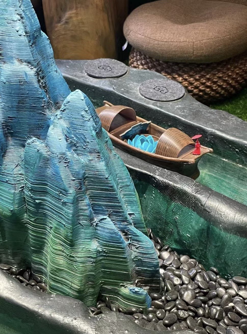 Miniature mountain landscape with a small wooden boat and red figurine on a water feature, surrounded by rocks, with two round stone stepping stones and a cushioned stool in the background.
