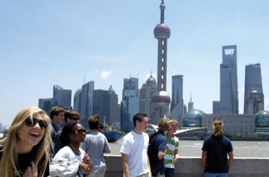 Group of tourists standing on a waterfront promenade with Shanghai skyline, including the Oriental Pearl Tower, in the background.