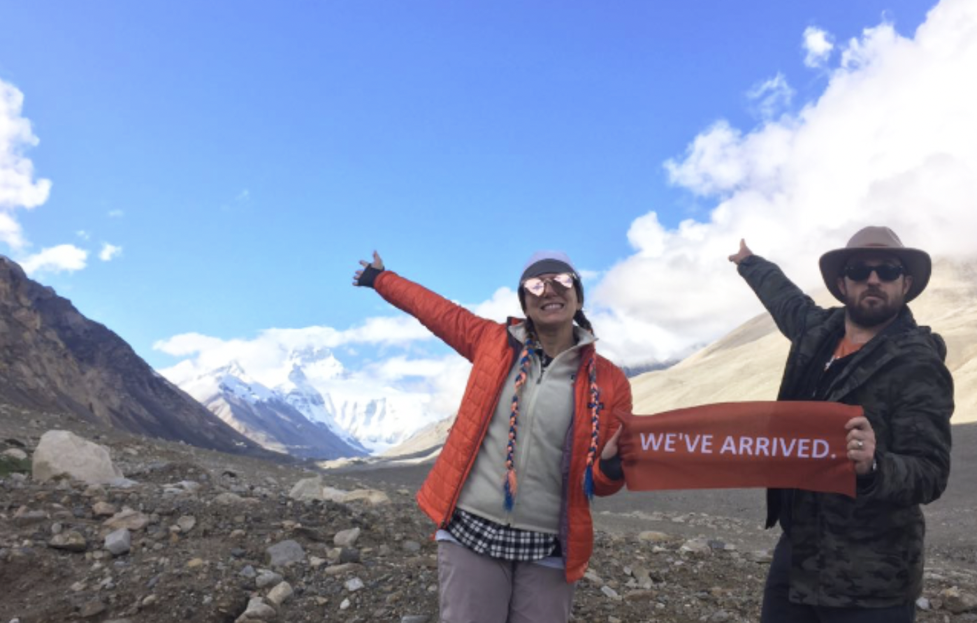Two smiling hikers, a woman and a man, standing on rocky terrain in a mountainous area, holding a red banner that says 'WE'VE ARRIVED.' The woman is wearing sunglasses, a gray beanie, a red jacket, and a plaid shirt, while the man is wearing a wide-brimmed hat, sunglasses, and a dark jacket. They are pointing towards snow-capped mountains and a partly cloudy sky in the background.