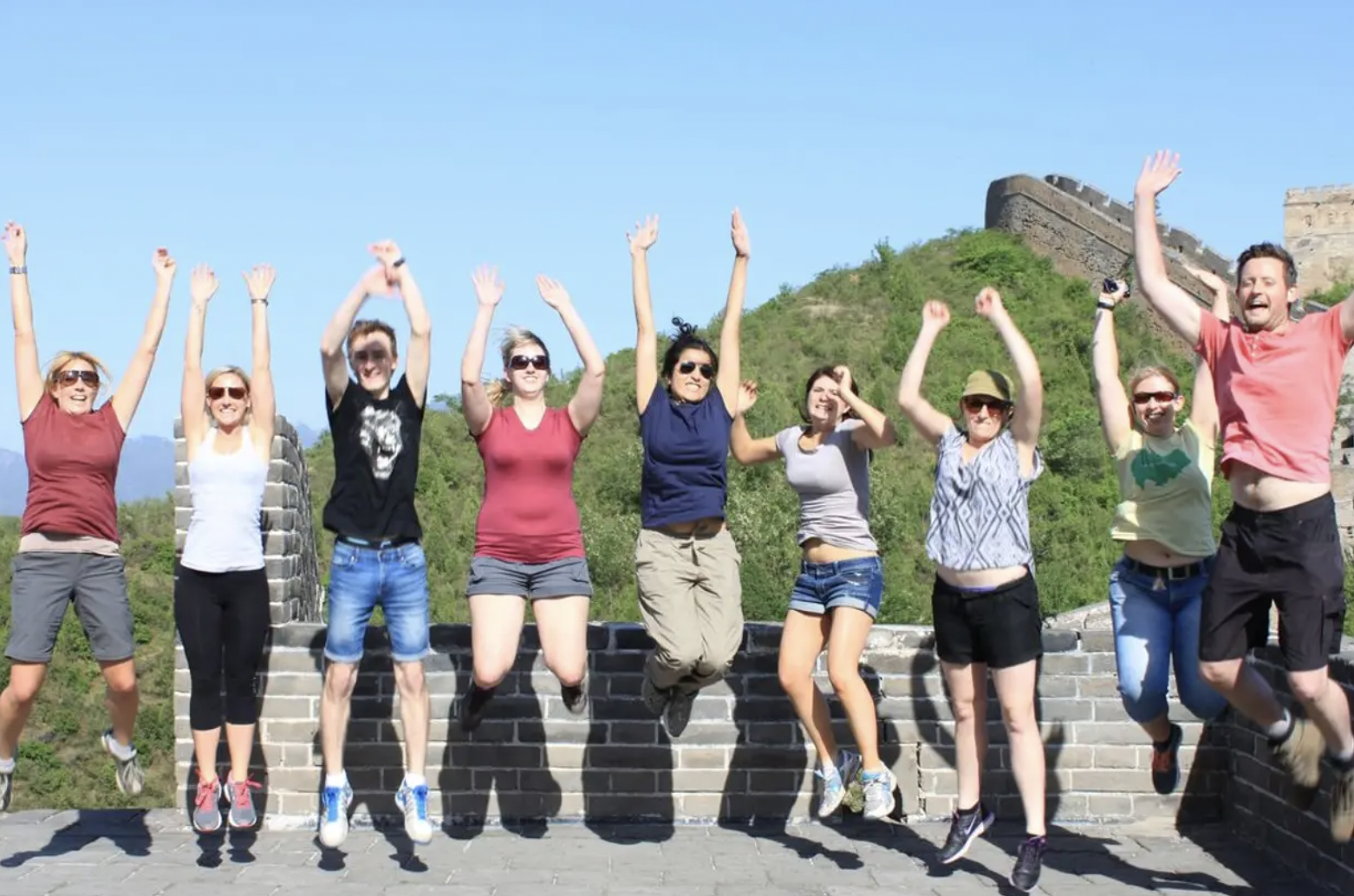Group of people jumping and smiling on the Great Wall of China with green hills and the wall in the background.