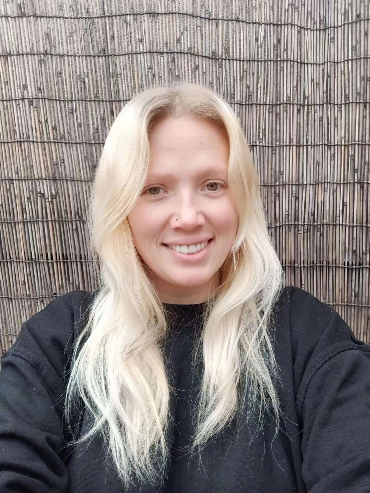 A smiling young woman with long blonde hair, wearing a black sweatshirt, standing in front of a bamboo fence.
