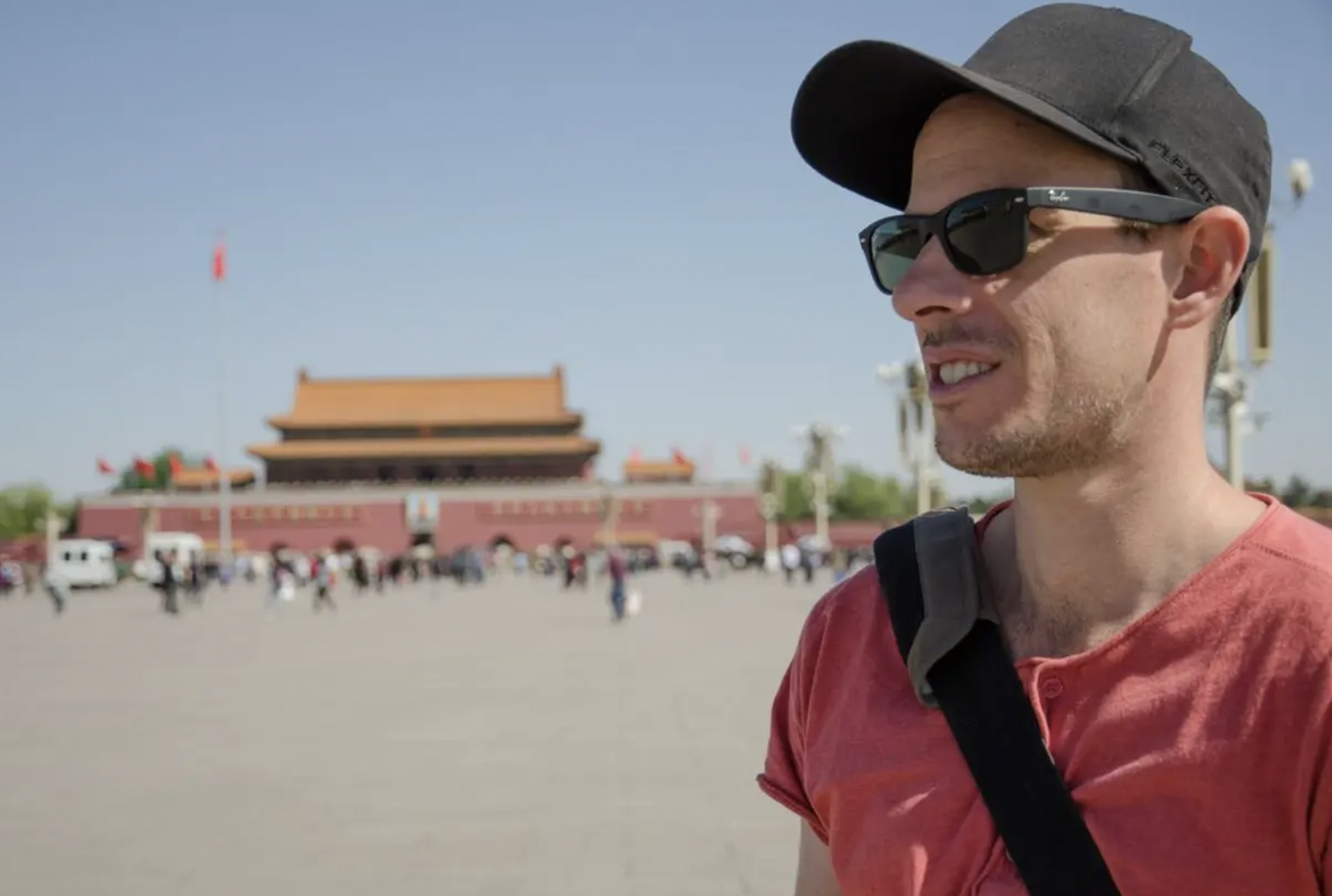 A man wearing sunglasses, a black cap, and a red shirt stands outdoors near a large traditional Asian-style building with a yellow roof, in front of a crowd and a wide open space under a clear sky.