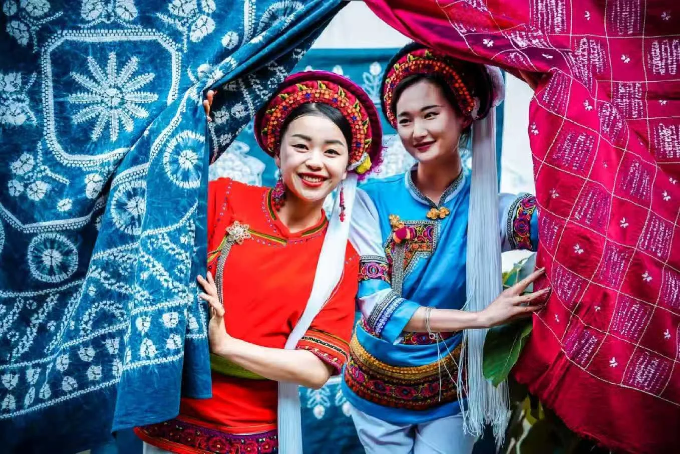 Two women in colorful traditional costumes and headpieces smiling and holding up vibrant textile fabrics.