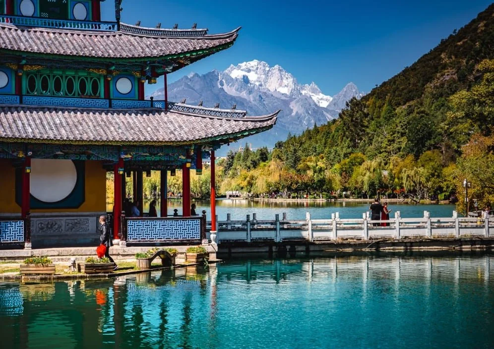 Traditional Chinese pavilion beside a calm lake with mountains in the background and a wooded area with trees on the opposite side of the lake.