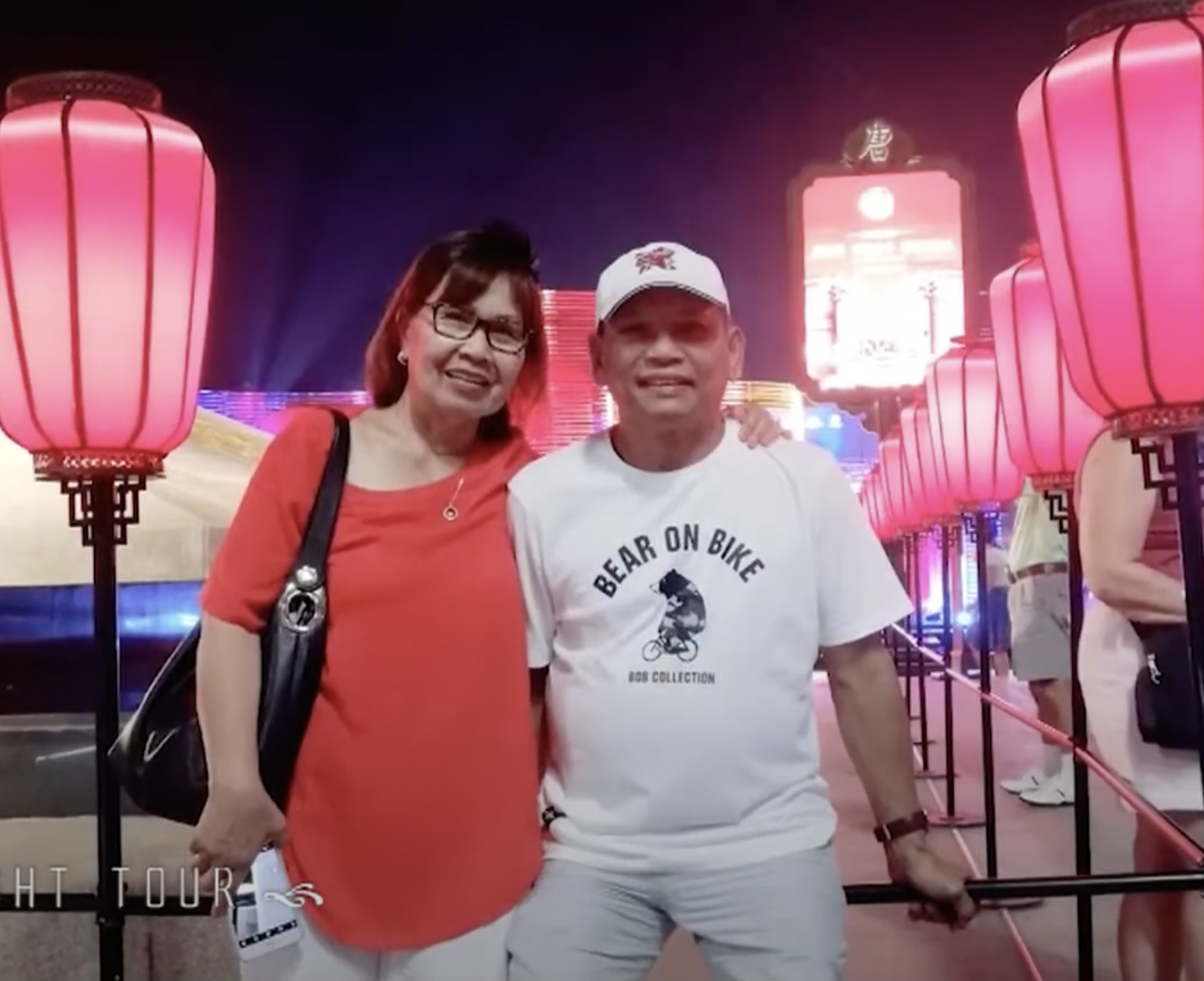 A woman and a man posing together at night with pink lanterns illuminated behind them.