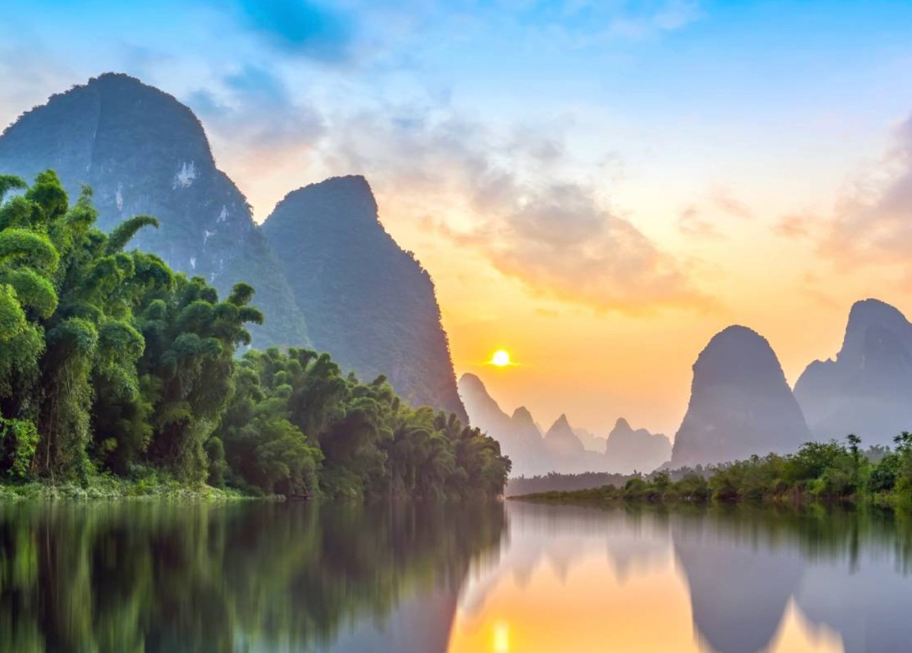 China Travel specialists Sunset over a river with karst mountains and lush greenery on either side, reflected in the water.