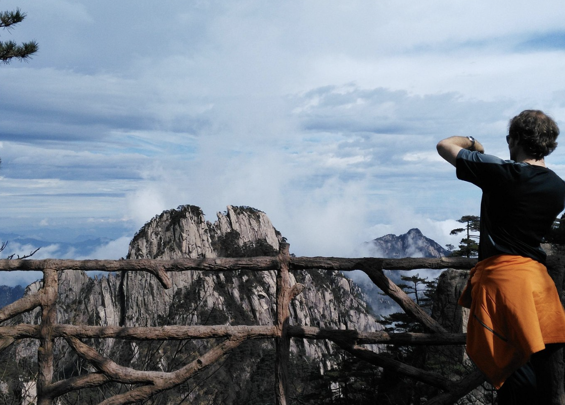 A person standing on a viewing platform made of logs, overlooking mountainous terrain with rugged peaks and a dramatic sky.