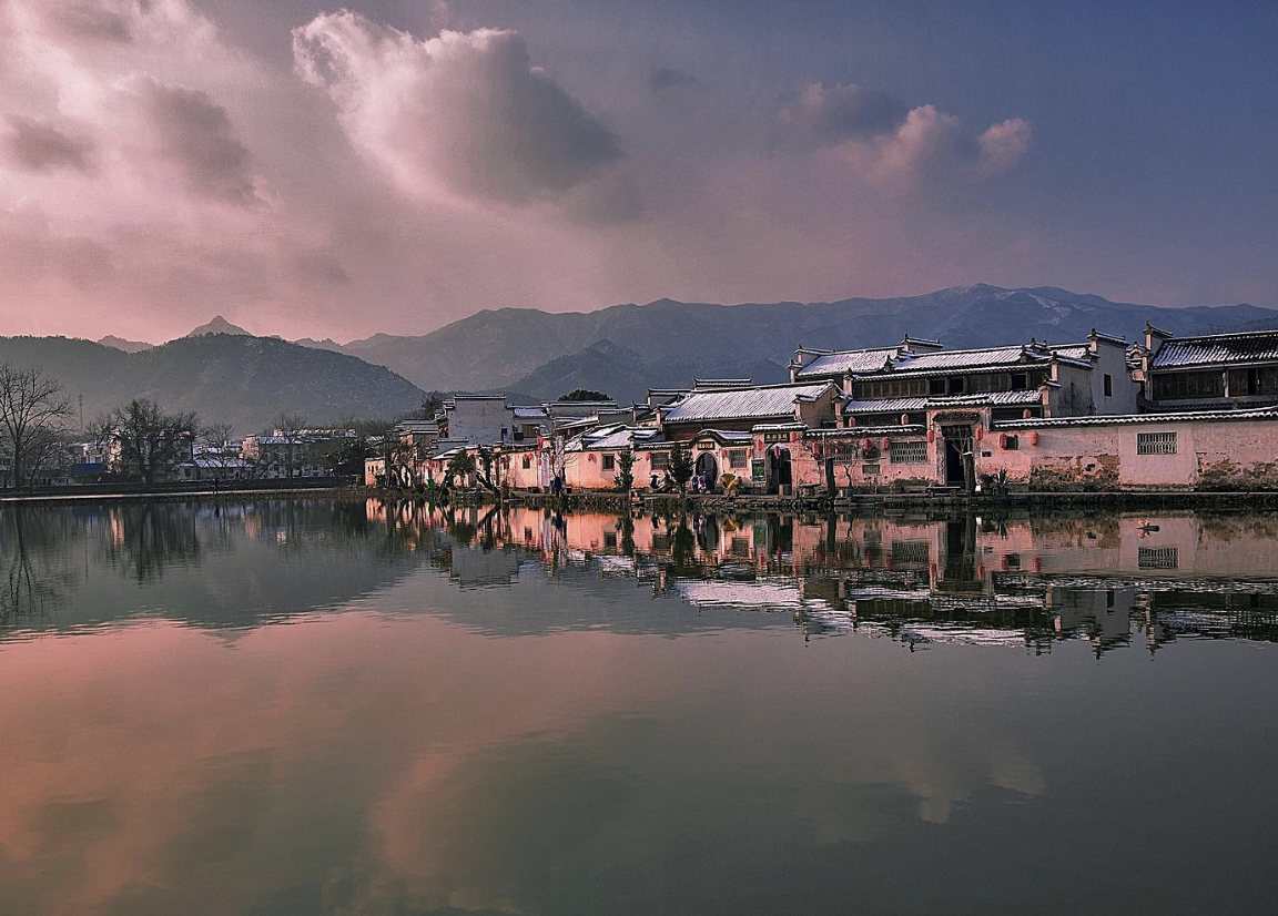 Traditional Chinese buildings along a river with mountains in the background at sunset