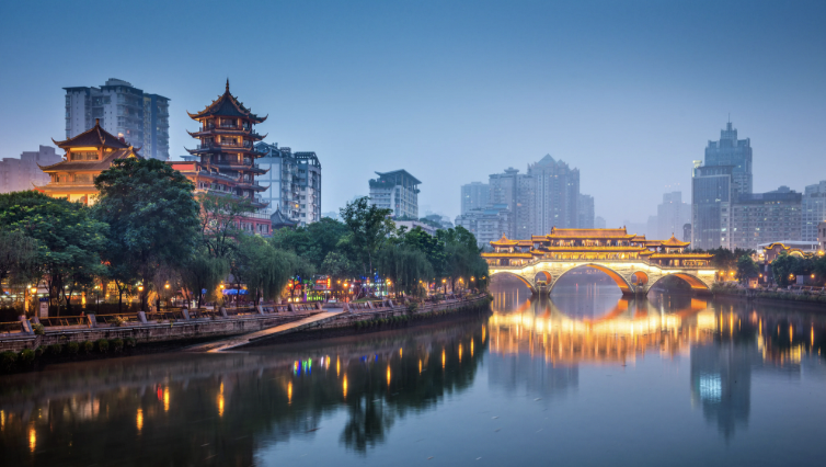 Cityscape with traditional Chinese temple architecture, modern high-rise buildings, and a bridge over a river, illuminated at dusk.