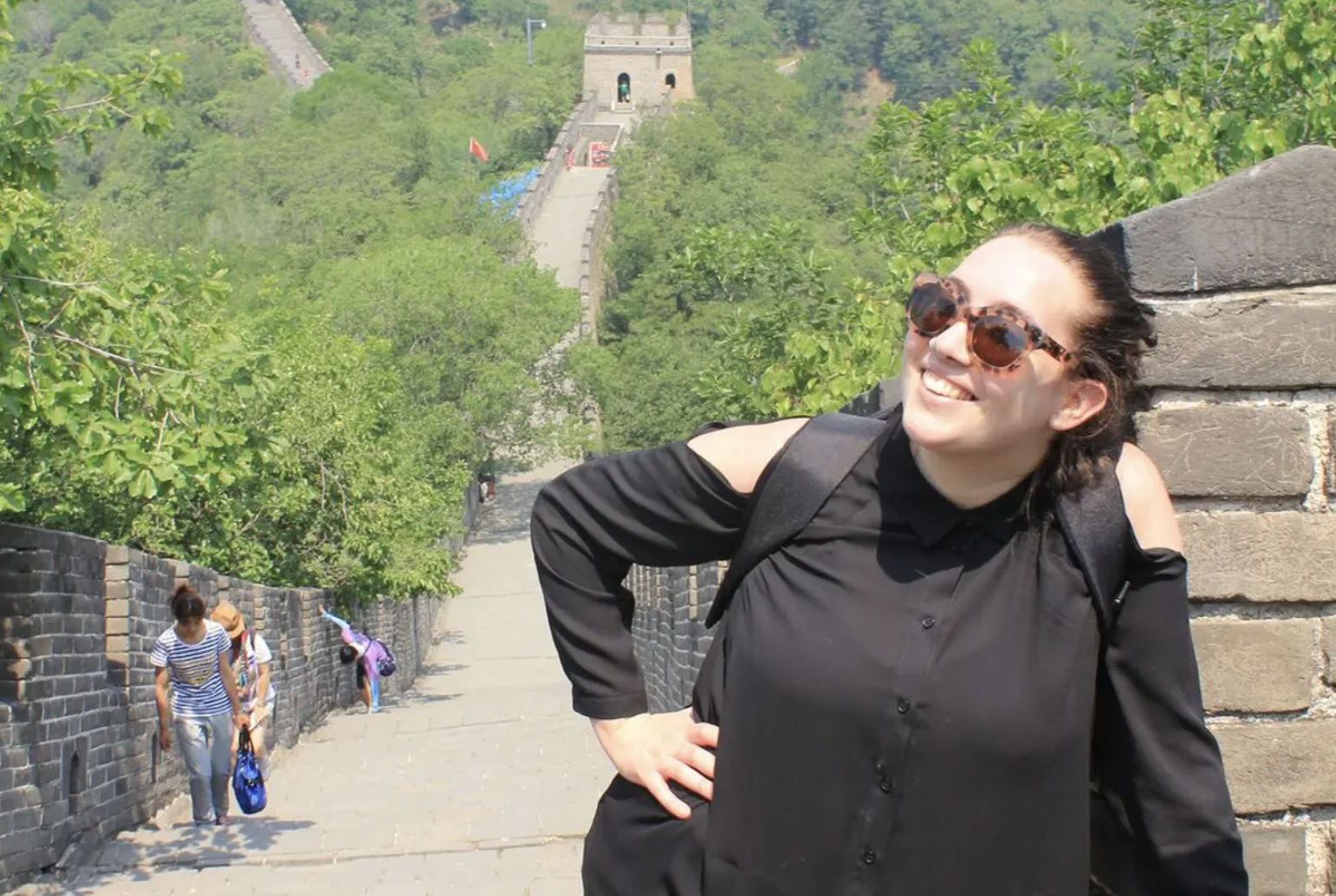 A smiling woman wearing sunglasses and a black shirt leaning against a stone wall on a historic stone wall with a staircase leading up to a watchtower in the distance. There are green trees and a few other people walking up the wall.